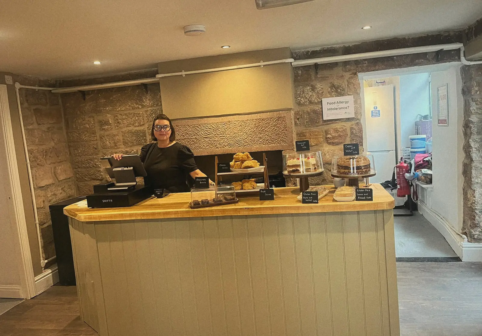 Amy behind the counter at Amy's Coffee Shop, with stone walls and cakes on display